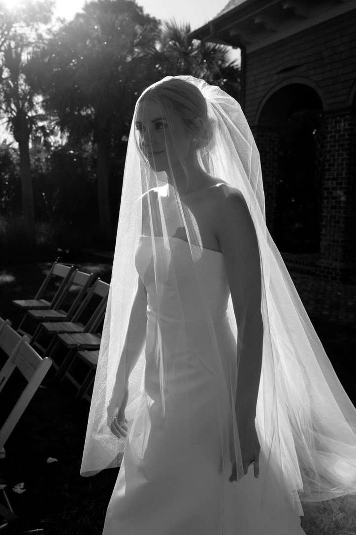 Black and white photo of a bride in a wedding dress and veil outdoors, with chairs lined up behind her and trees in the background.