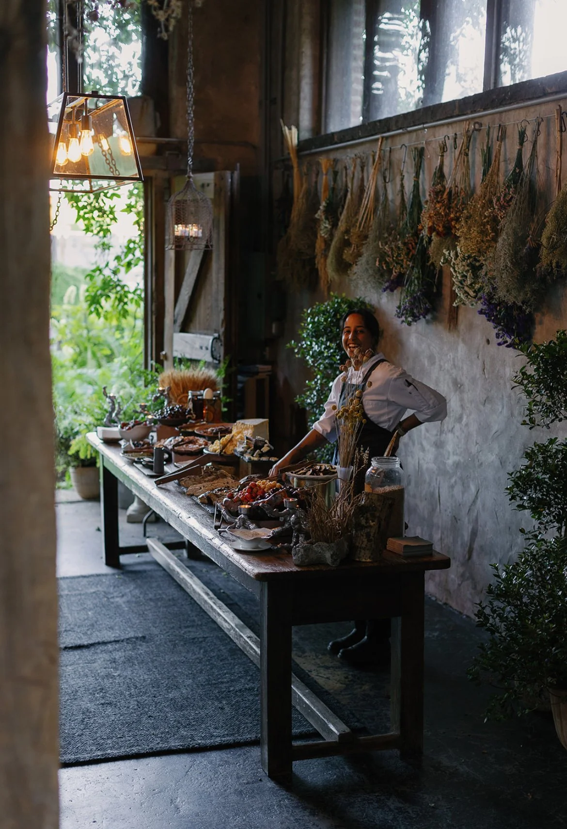 A smiling woman in a white shirt and black apron standing behind a rustic wooden table filled with various food items, inside a cozy, humidity with dried flowers hanging on the wall and greenery outside through an open door.