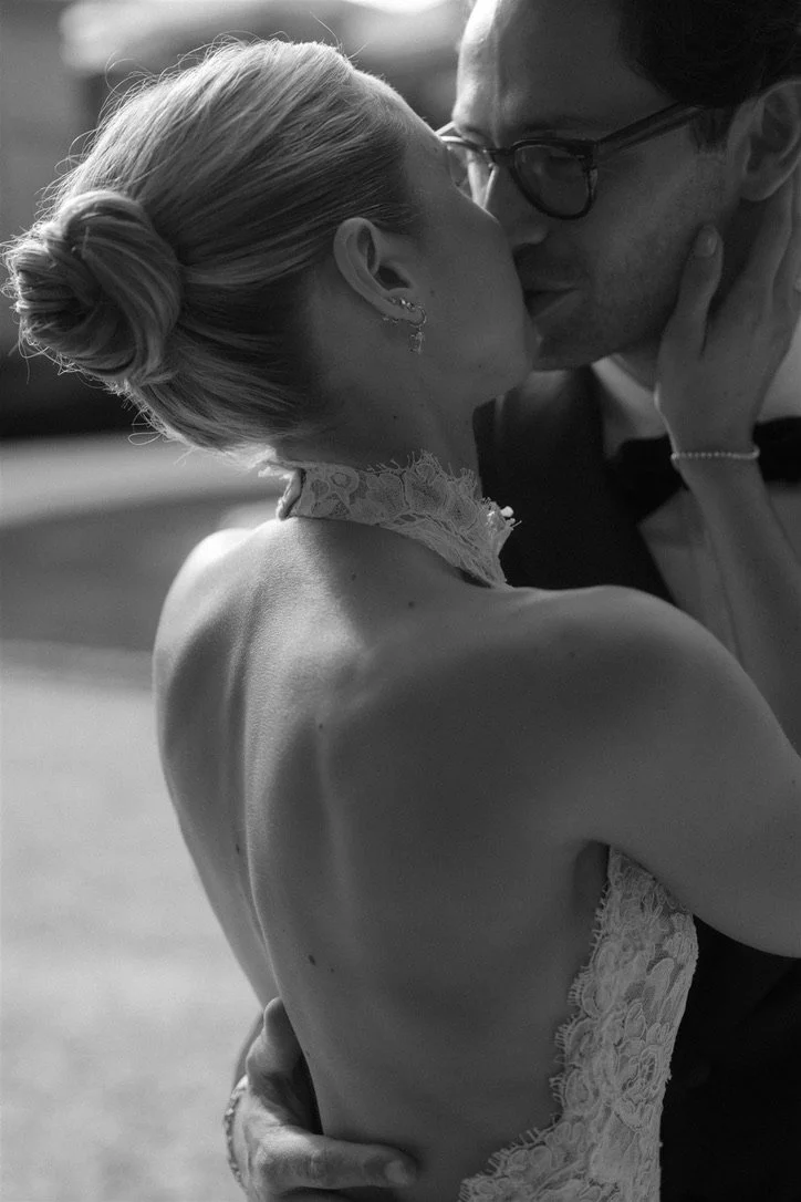 A black and white photo of a couple kissing, with the woman wearing a lace dress and the man wearing glasses and a tuxedo.