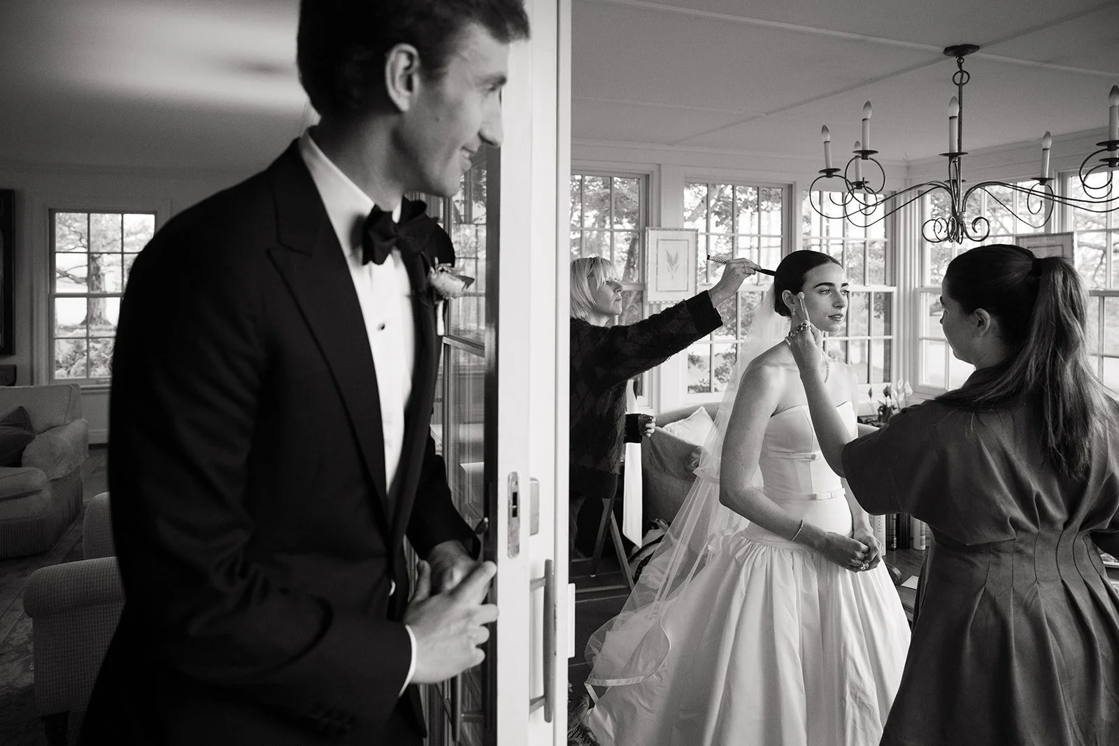 A bride sitting in a room while a makeup artist applies makeup, with a woman in the background doing her hair, and a man in a tuxedo standing in the doorway smiling.