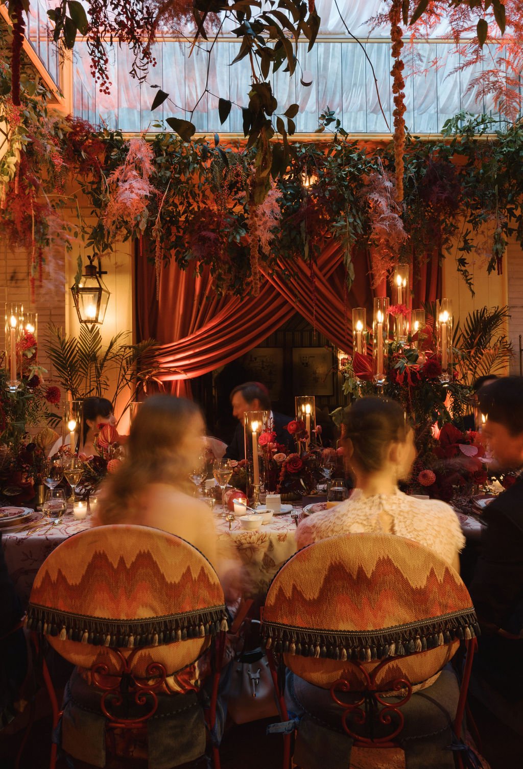A decorated indoor event space with a high ceiling, hanging greenery, and draped red curtains. The space features warm lighting, candles, and floral arrangements on a dining table where elegantly dressed people are seated, enjoying a meal.