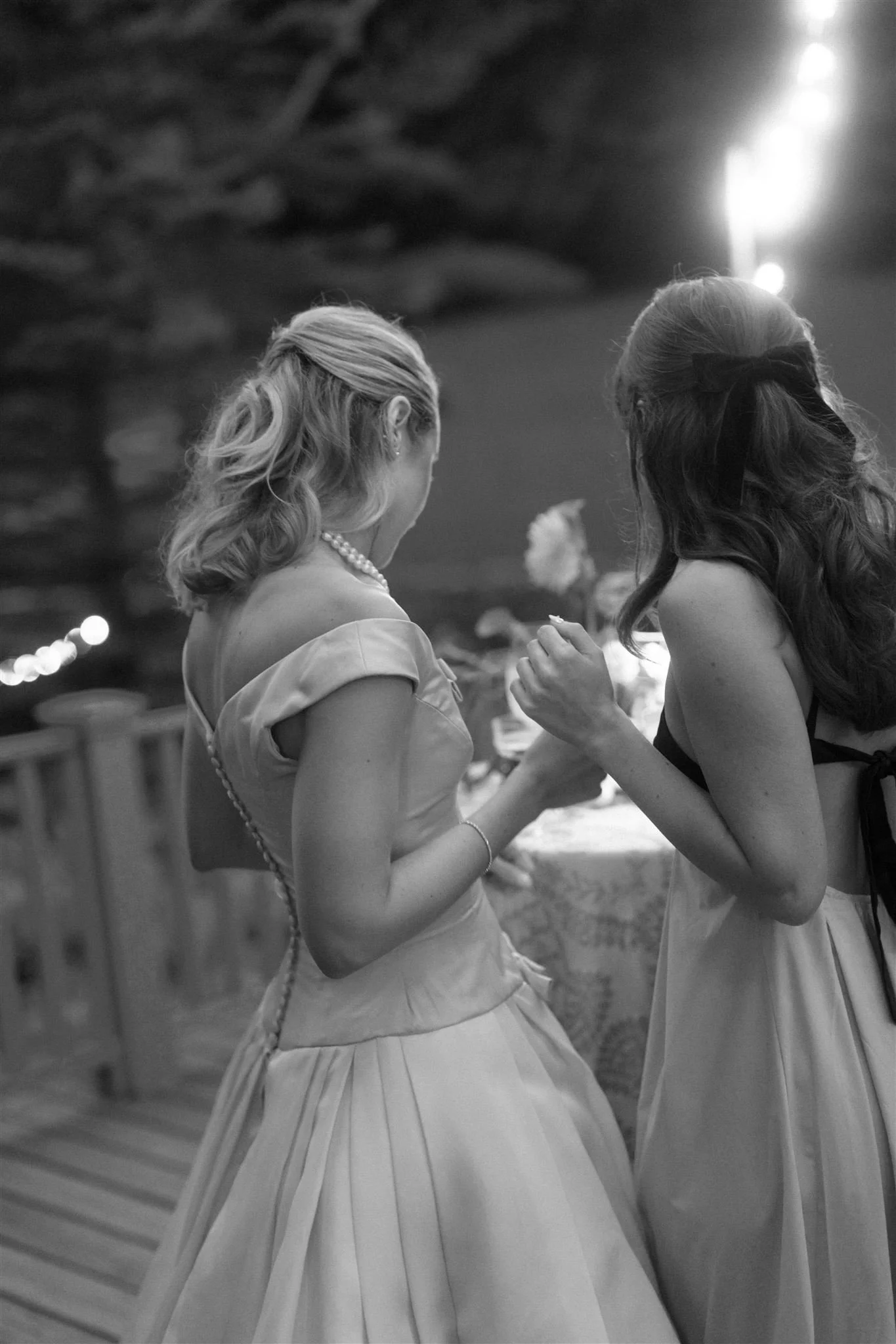 Two women sharing a moment, one in a vintage style dress with jewelry and the other in a dress with a ribbon in her hair, in a warmly lit indoor setting.