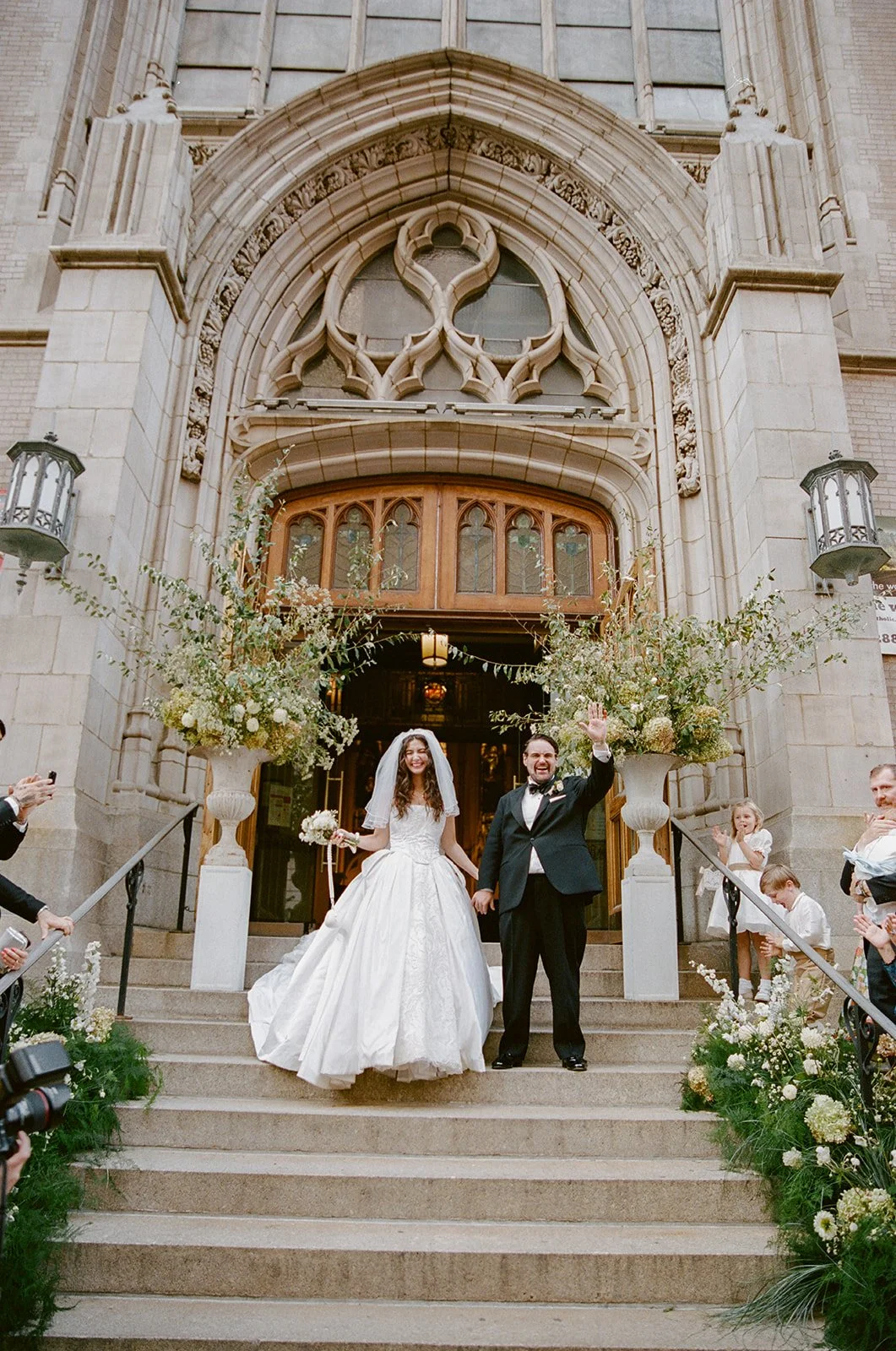A newlywed couple standing on the steps of a church, celebrating their wedding with friends and family. The bride is in a white wedding gown holding a bouquet, smiling, and the groom is in a black tuxedo waving. There are guests on each side clapping