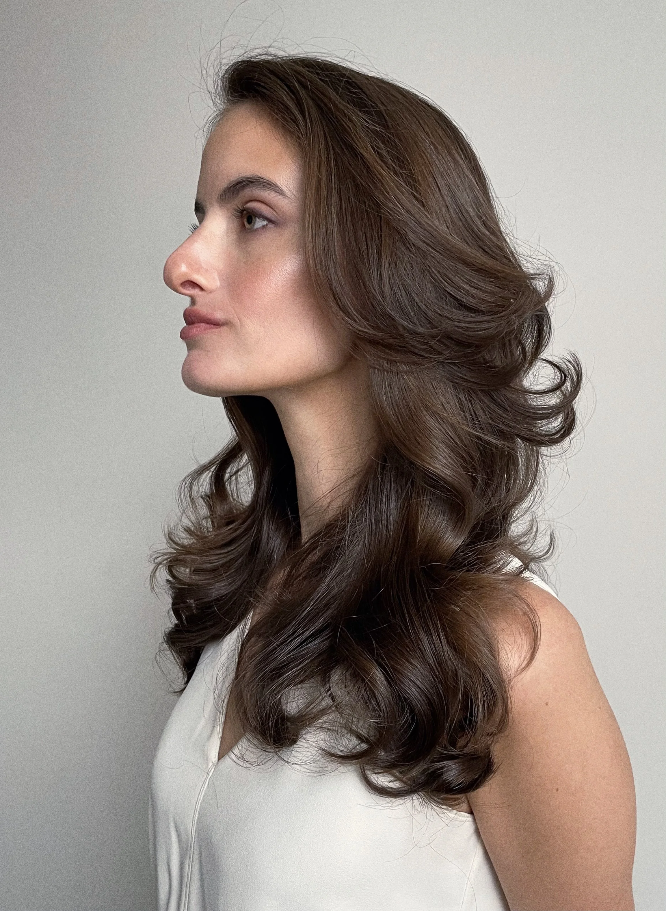 Side profile of a woman with long, wavy brown hair, wearing a white sleeveless top, against a plain light background.