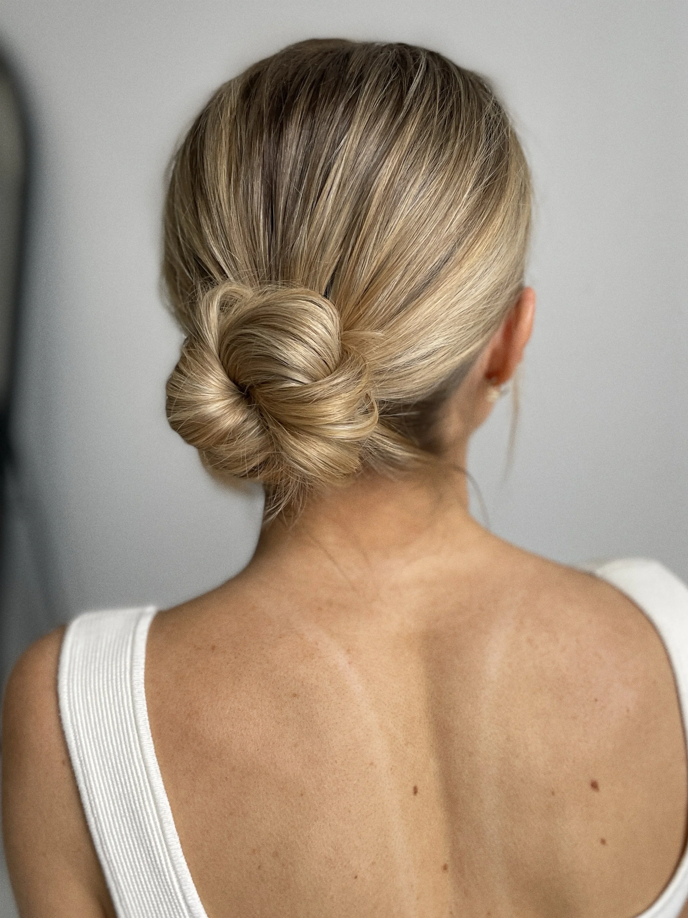 Back view of a woman with blonde hair styled in a twisted bun, wearing a white sleeveless top.