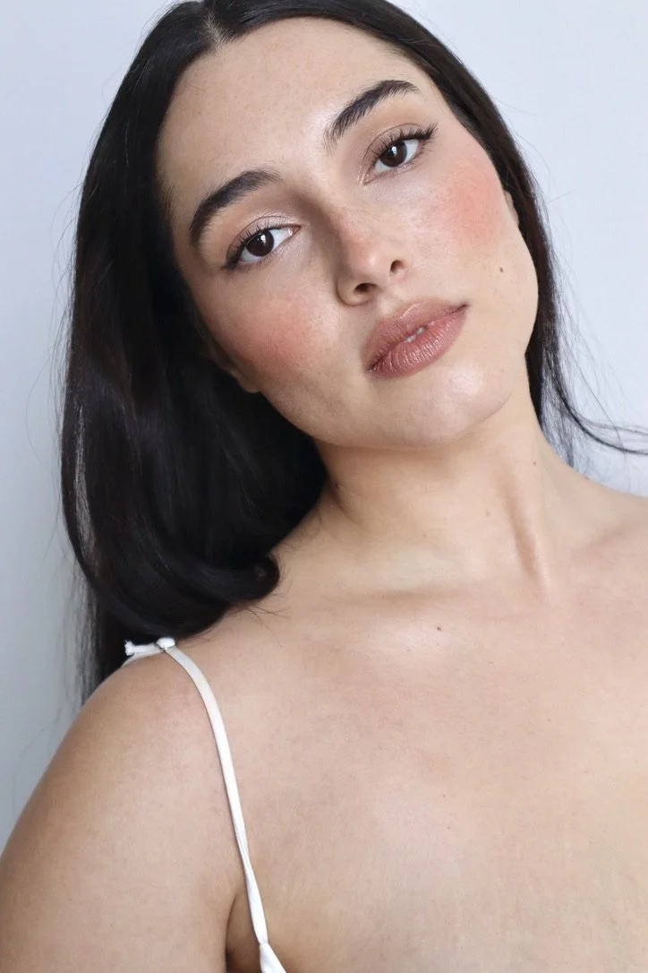 A close-up of a young woman with long dark hair and light skin, looking at the camera with a neutral expression, wearing a thin white strap top.