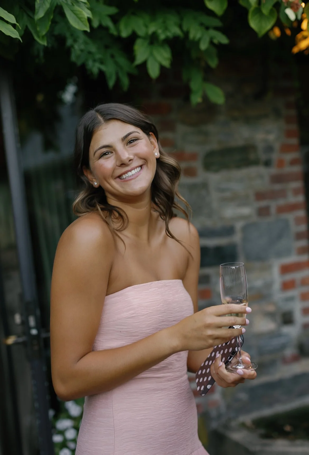 A young woman in a strapless pink dress holding a glass of champagne and smiling outdoors near a stone wall.