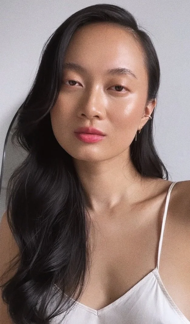 A young woman with long, wavy black hair wearing a white spaghetti strap top, looking directly at the camera with a neutral expression.