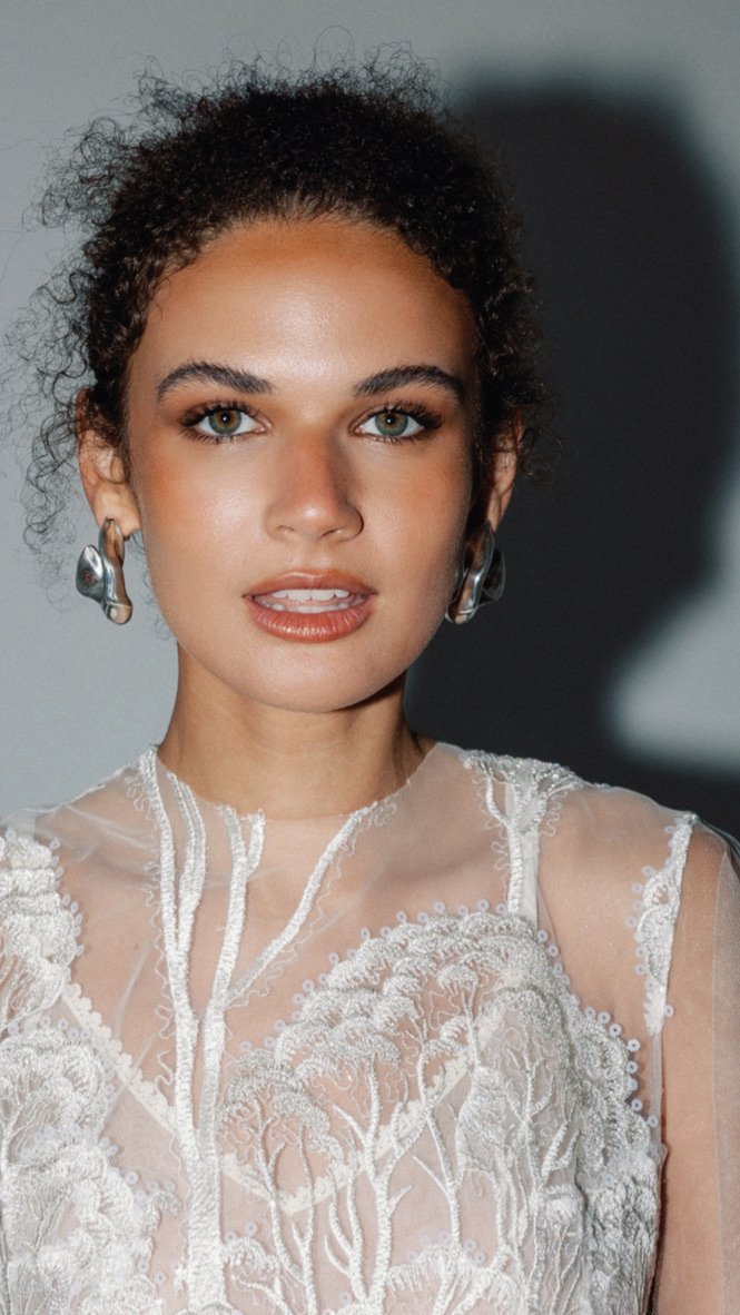 A young woman with curly hair, wearing bold silver earrings and a white lace top, looking directly at the camera.