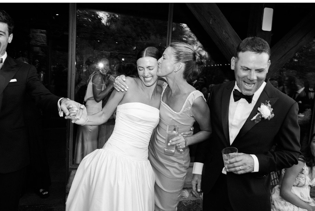 Black and white photo of people celebrating at a wedding reception, with a bride and she is embraced by a woman and holding a drink, and a man in a tuxedo also holding a drink, all smiling