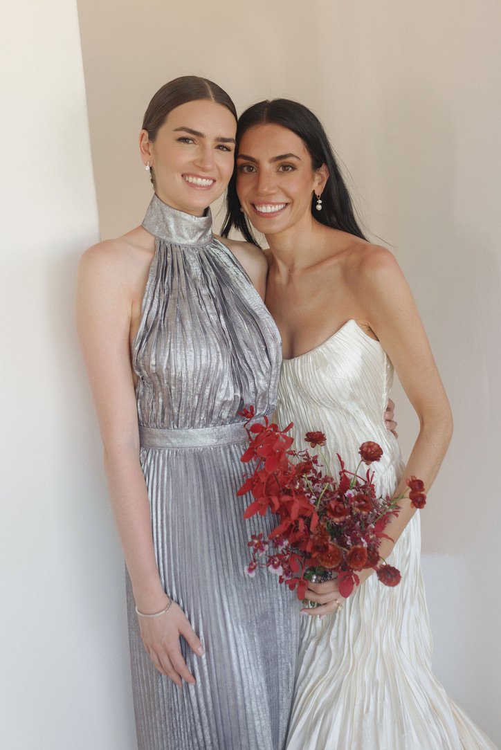 Two women smiling and standing close together, one holding a red flower bouquet, dressed in elegant outfits.