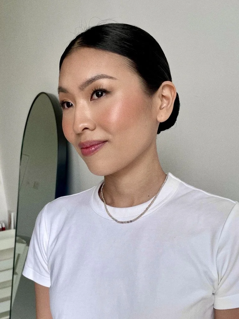 A woman with black hair styled in a bun, wearing a white t-shirt, silver chain necklace, and makeup, standing in front of a mirror.