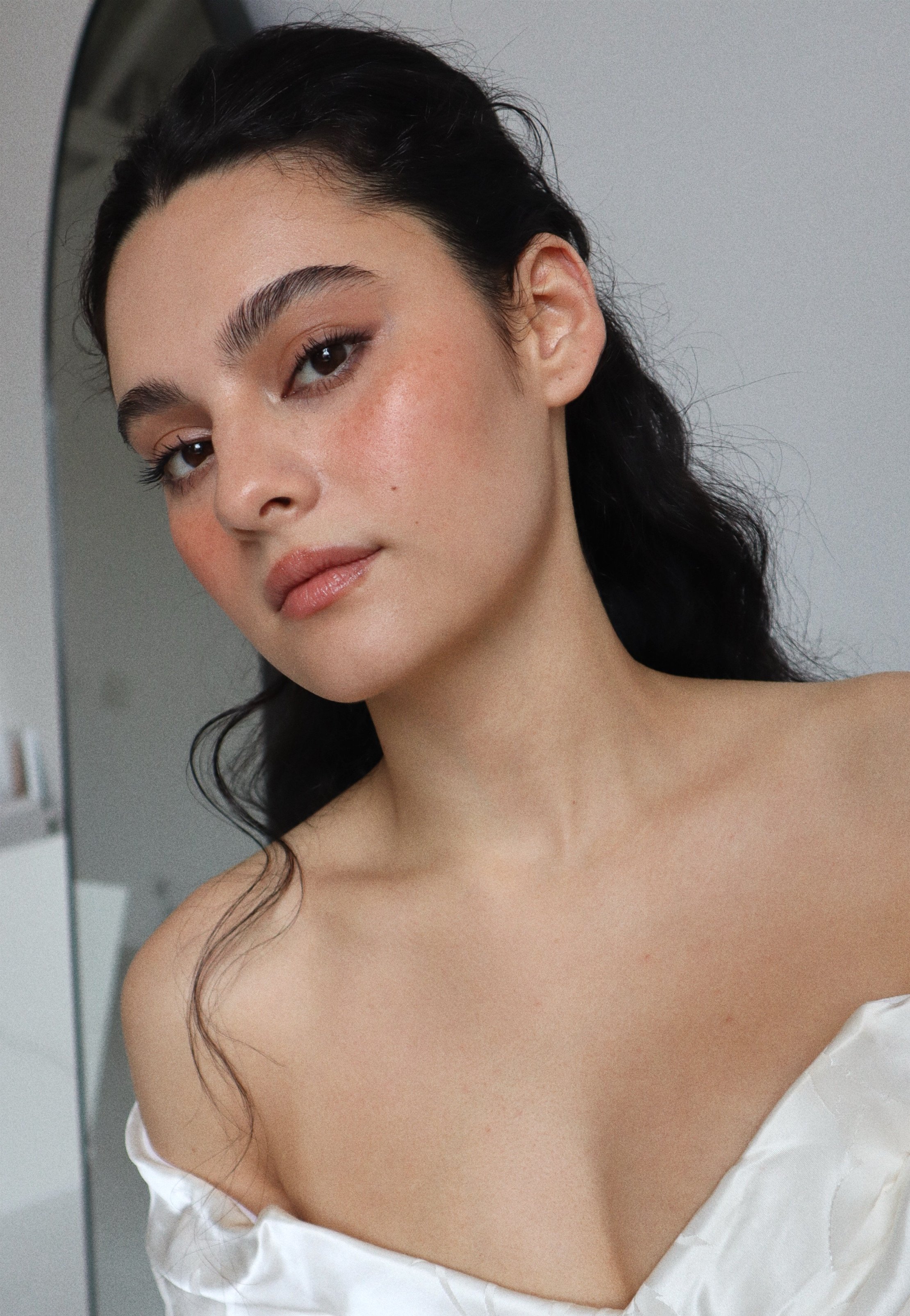 A young woman with long dark wavy hair, makeup, and a white off-the-shoulder top, looking at the camera in a close-up portrait.