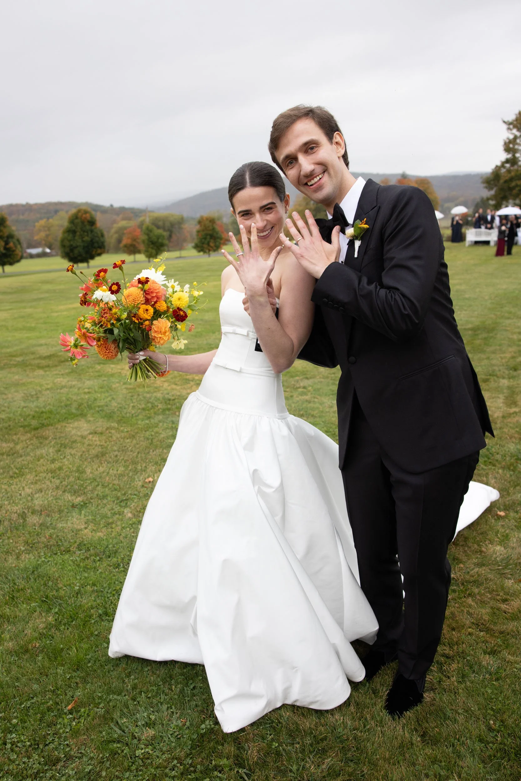 A newlywed couple outdoors on their wedding day, showing off their rings and holding a colorful bouquet of flowers, smiling happily.