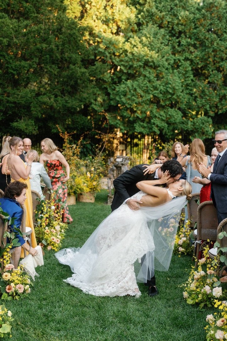 A bride and groom kiss during their outdoor wedding ceremony, surrounded by friends and family, with lush green trees and floral decorations.