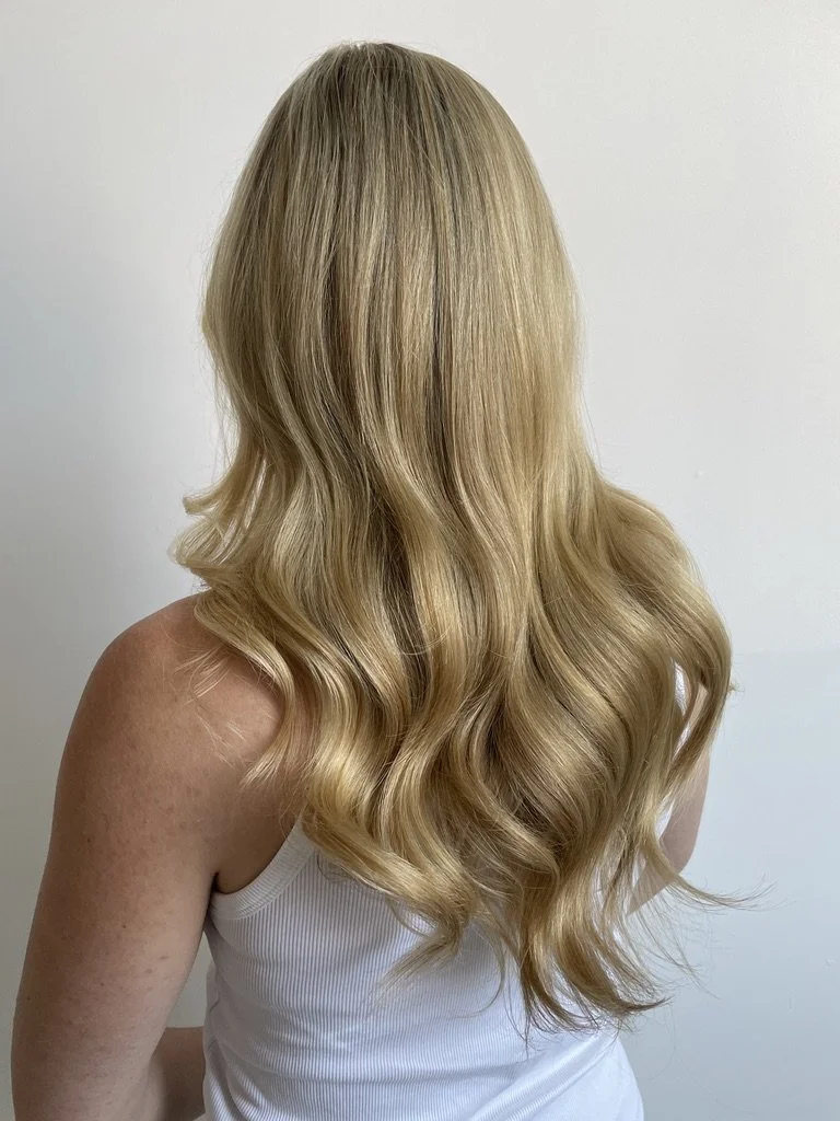 A woman with long, wavy blonde hair wearing a white sleeveless top, standing against a plain light-colored wall.