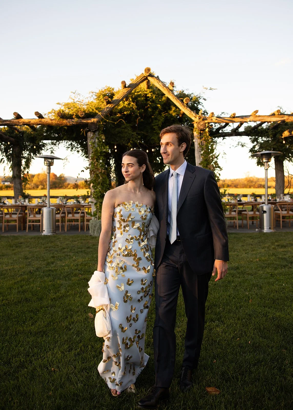 A newlywed couple walking outdoors in a garden during sunset. The bride is wearing a strapless white gown adorned with gold butterflies, holding a matching white clutch. The groom is in a dark suit with a light-colored tie. In the background, there i