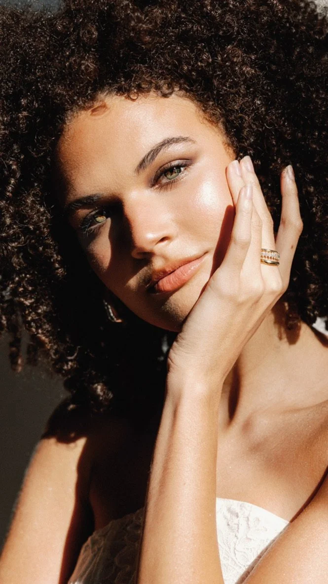 Close-up of a woman with curly hair, posing with her hand on her face, wearing a ring, and looking at the camera with a confident expression, illuminated by sunlight.