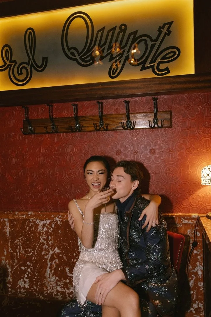 A young woman and a young man sitting closely together in a cozy, dimly-lit restaurant or bar, smiling and holding drinks, with a large yellow sign reading 'El Quintote' above them.
