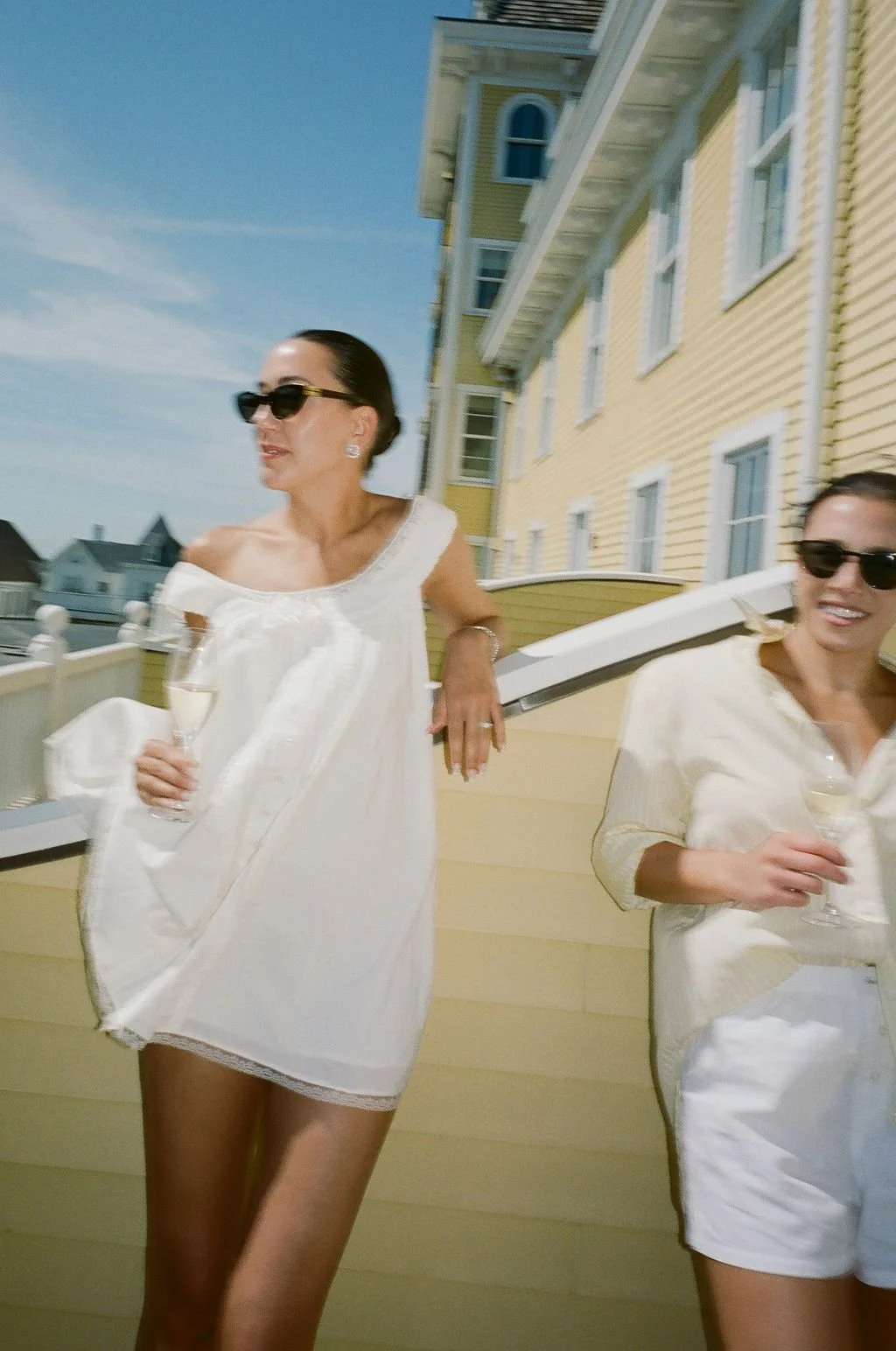 Two women wearing white dresses and sunglasses, holding glasses of champagne, on a balcony with a yellow house in the background.