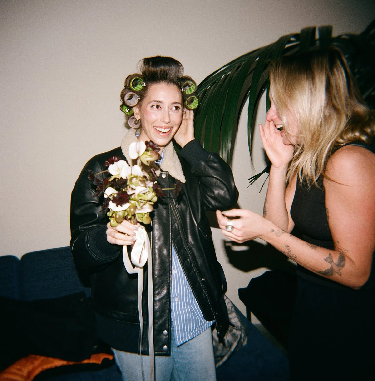 Two women laughing and sharing a moment indoors, one woman with hair in rollers holding a bouquet of flowers, the other woman with tattoos on her arm and blonde hair, covering her mouth while reacting to the woman with rollers.