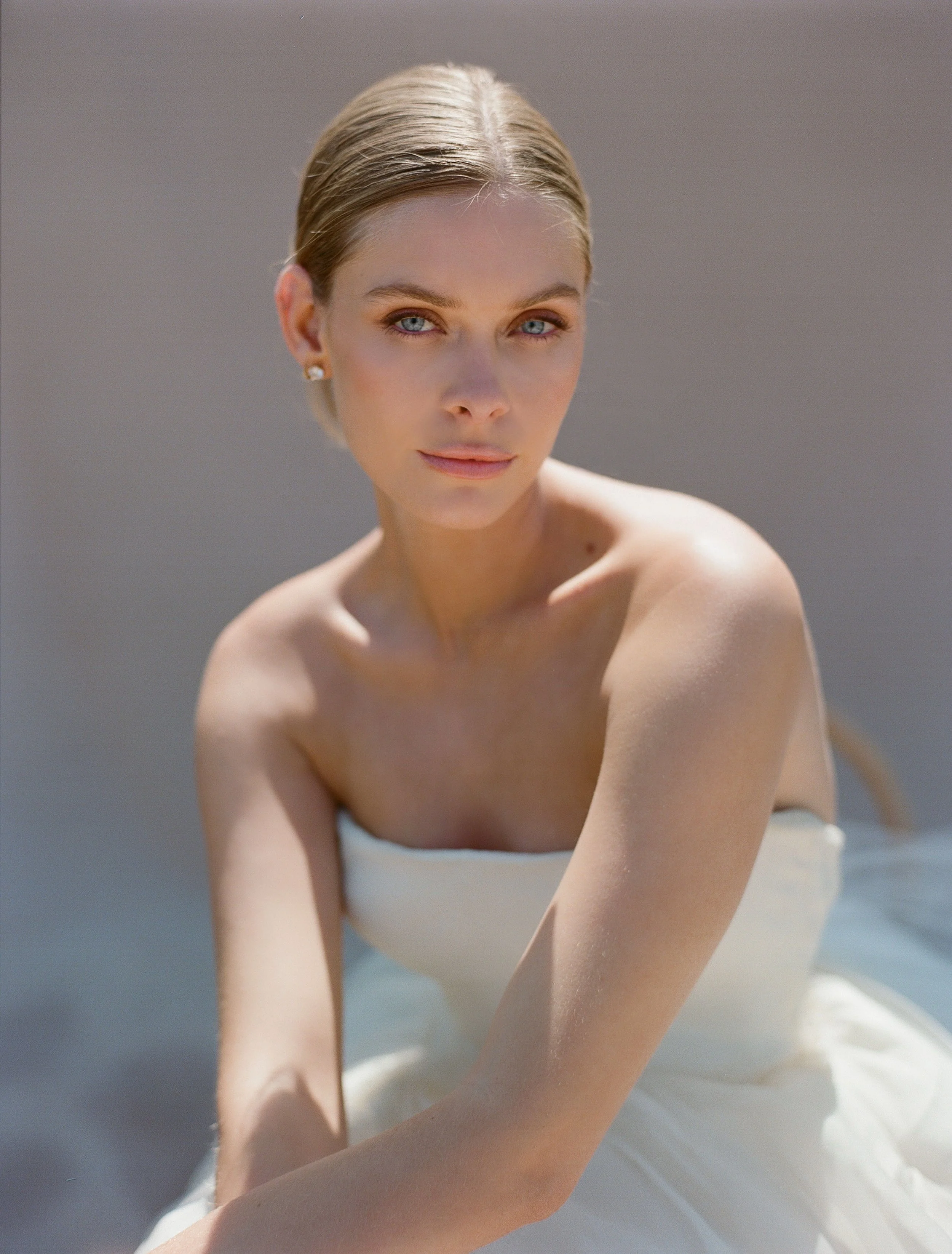 A woman with blonde hair and blue eyes looking at the camera, wearing a strapless white dress, with a neutral background.