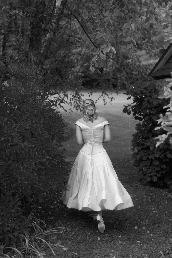 Woman in a wedding dress walking away through a garden path surrounded by trees and bushes.