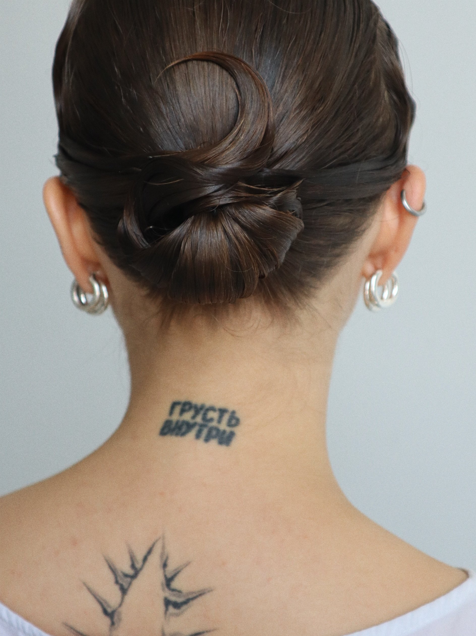 Back of a woman with a braided and twisted hairstyle, multiple earrings, a tattoo on the neck with text in a foreign language, and another tattoo on the upper back.