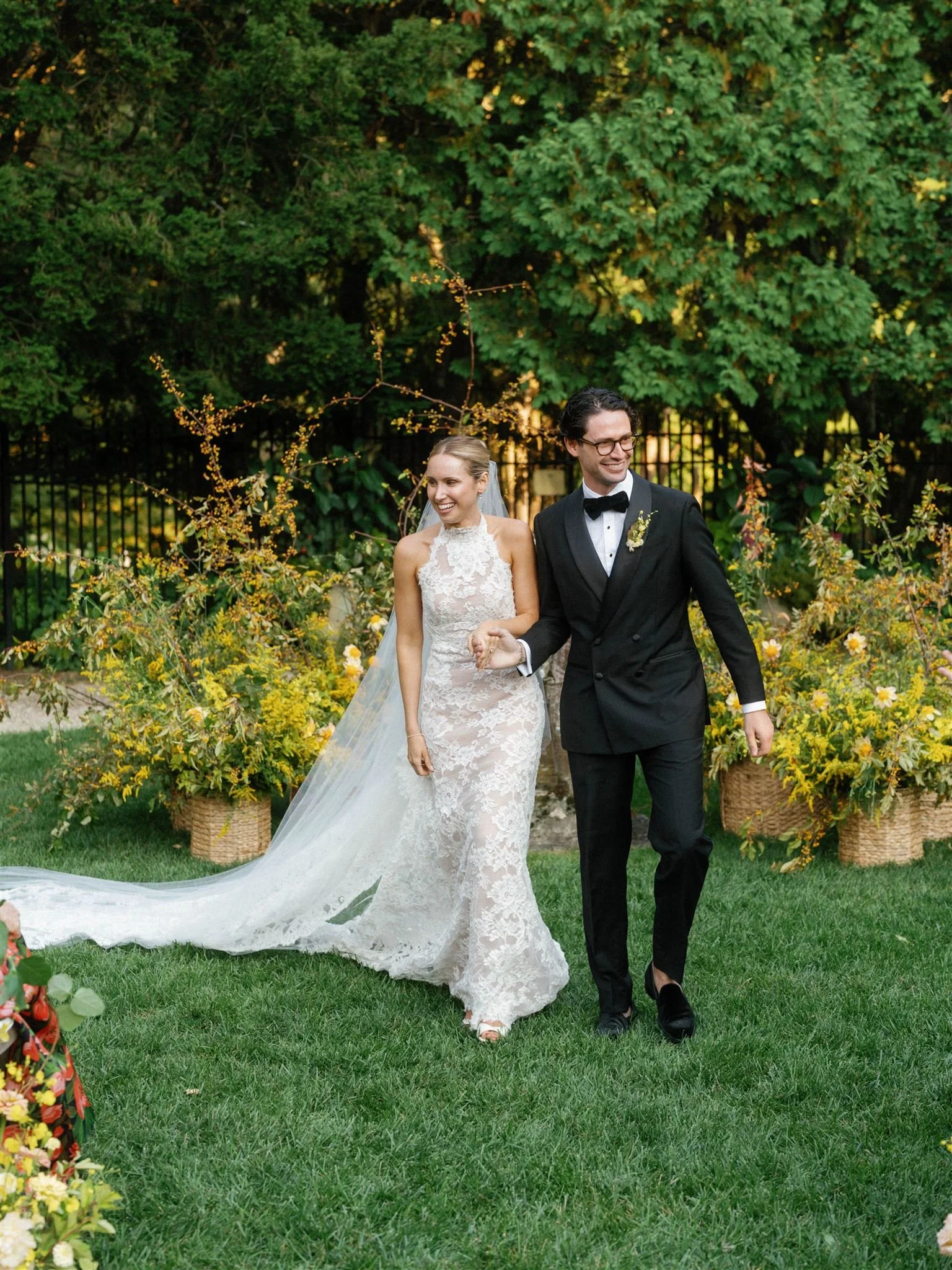 A bride and groom walking hand in hand outdoors during their wedding, surrounded by greenery and floral arrangements.