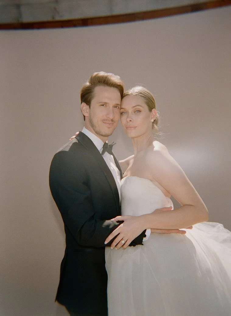 A man and a woman dressed in formal wedding attire posing together, with the man in a tuxedo and the woman in a strapless white wedding gown.