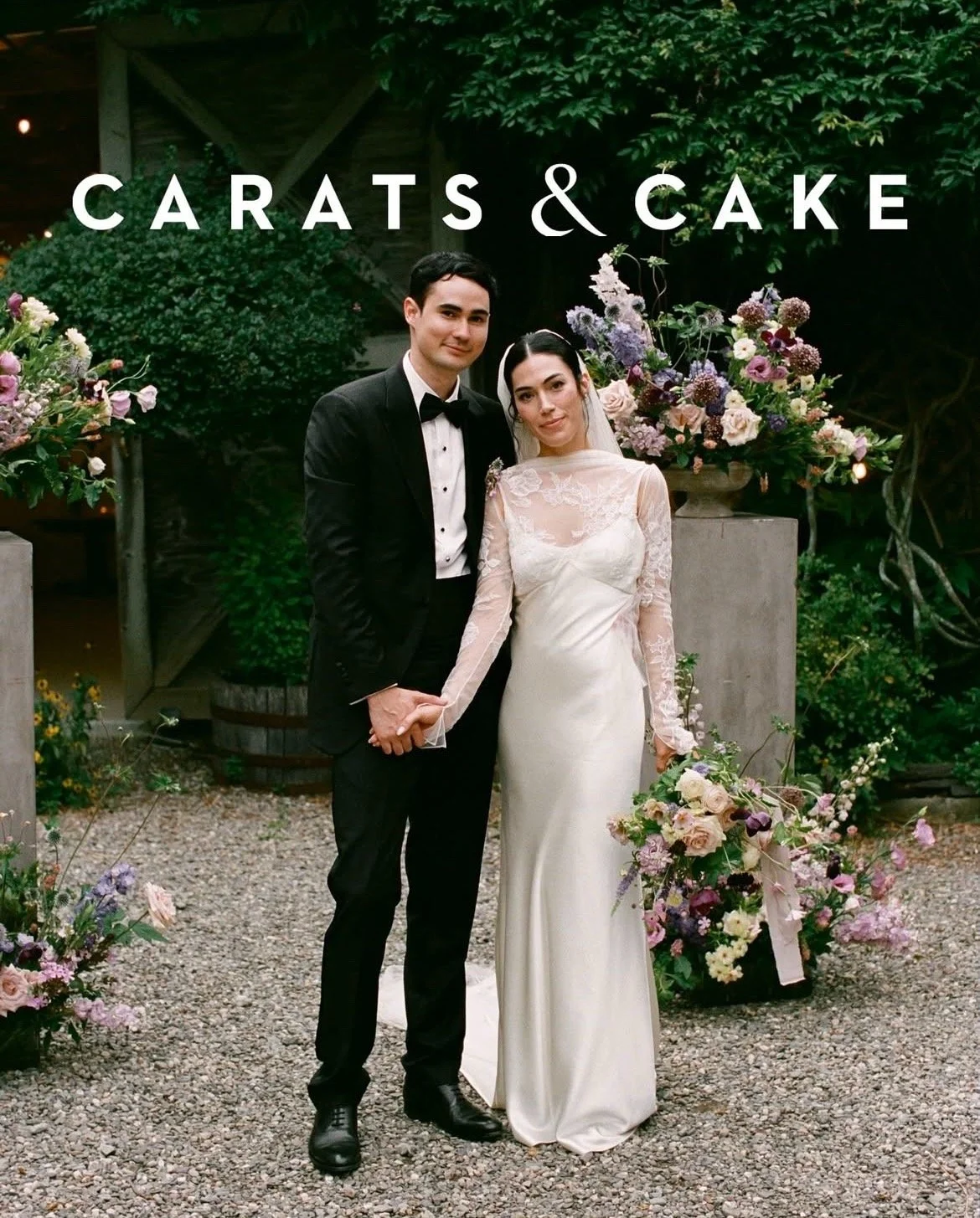 A bride and groom holding hands and posing for a wedding photo in front of floral arrangements and greenery.