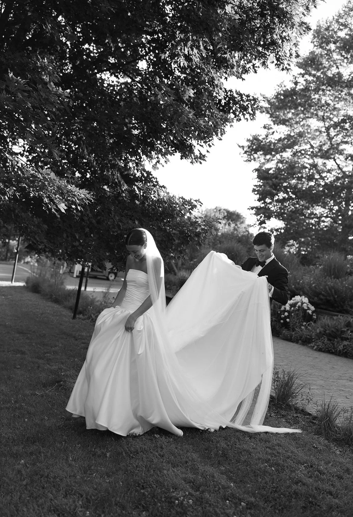 A bride in a strapless wedding gown with a long veil and a groom in a tuxedo standing outdoors on grass and a paved path, with trees and bushes in the background.