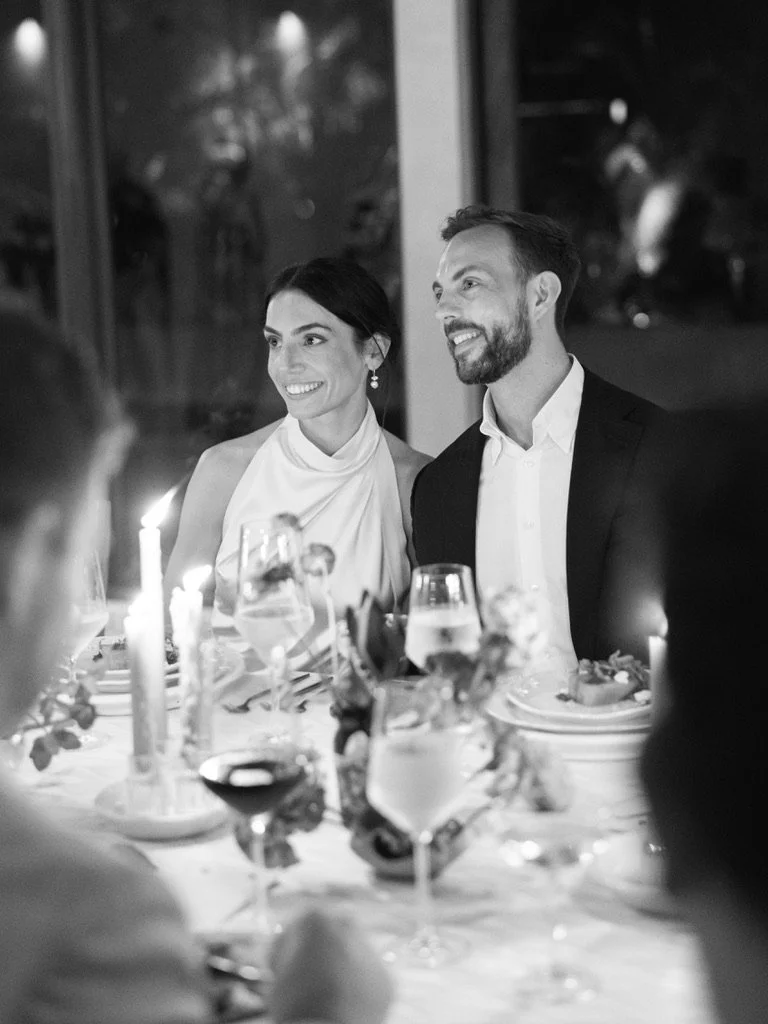 A smiling couple sits at a dinner table, dressed formally, in a restaurant or banquet setting with glasses, candles, and elegant tableware visible.