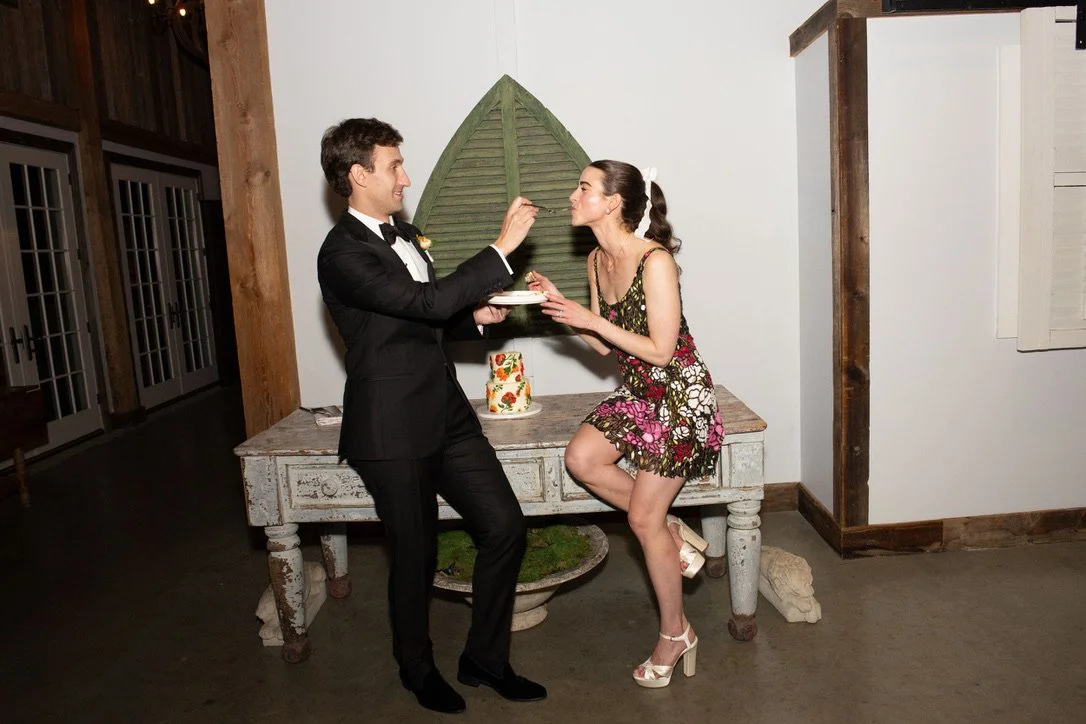 A woman in a floral dress and high heels playfully feeding a man in a tuxedo at a social gathering.