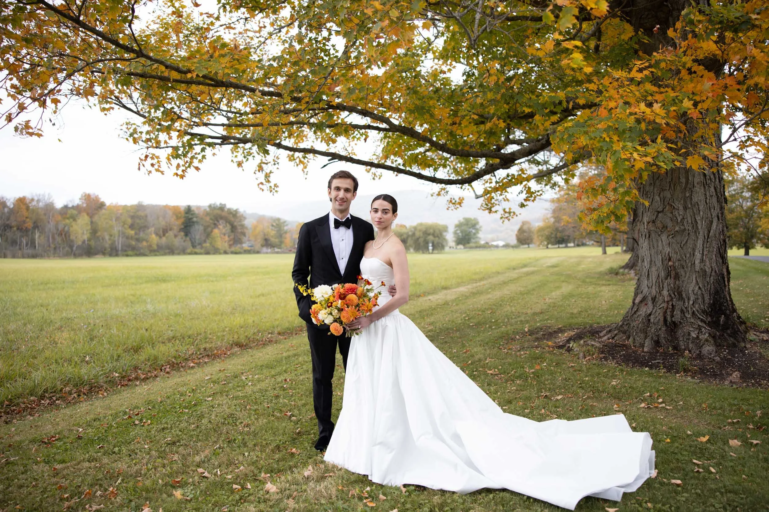 A newlywed couple standing together under a large tree in a wide open grassy field. The bride is wearing a white wedding dress and holding a colorful bouquet. The groom is dressed in a black tuxedo with a bow tie. The scene is set outdoors during fal