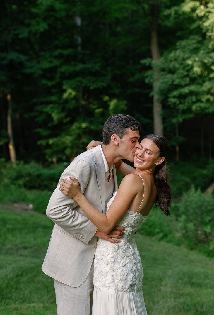 A happy bride and groom sharing a kiss outdoors in a forested area, with the groom in a light-colored suit and the bride in a white dress.