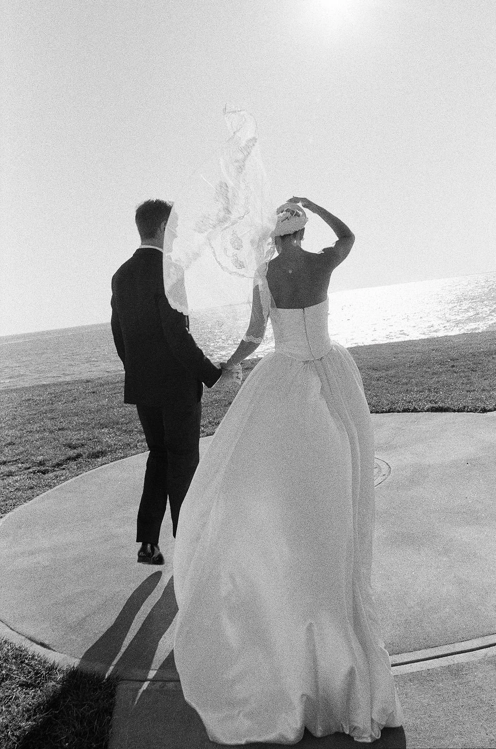 A black and white photo of a bride and groom standing outdoors near the water, holding hands, with the bride wearing a long gown and the groom in a suit, and the bride holding a hat to her head.