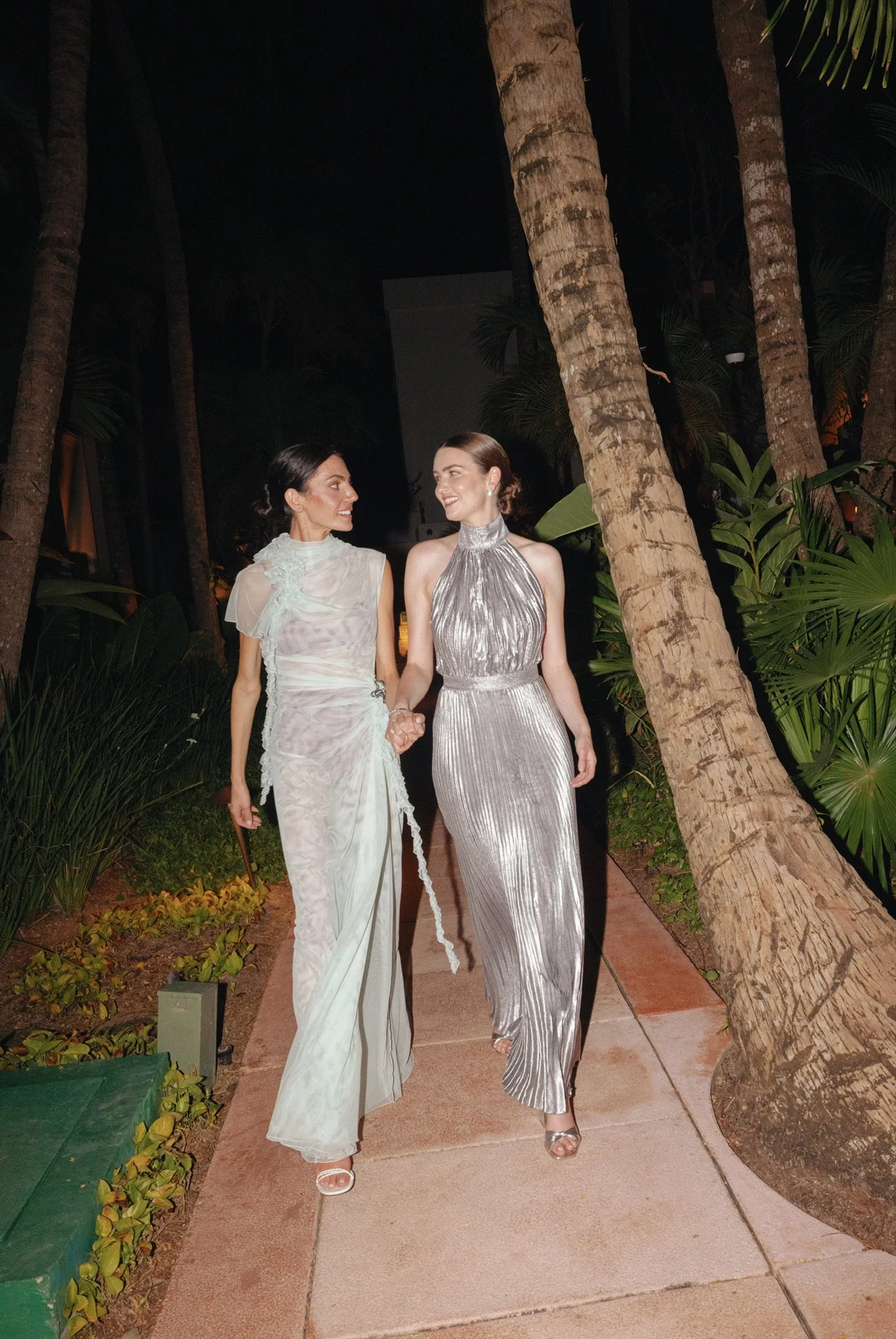 Two women holding hands and walking along a sidewalk at night, surrounded by tropical plants and palm trees, dressed in elegant evening gowns, smiling at each other.