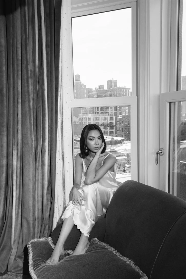 A woman sitting on a sofa by a large window with city buildings in the background, wearing a slip dress.