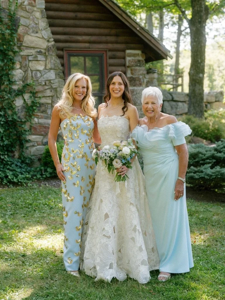 Three women standing outdoors in front of a stone and wood house, smiling, with trees in the background. The woman in the middle is wearing a wedding dress and holding a bouquet, suggesting a wedding celebration.