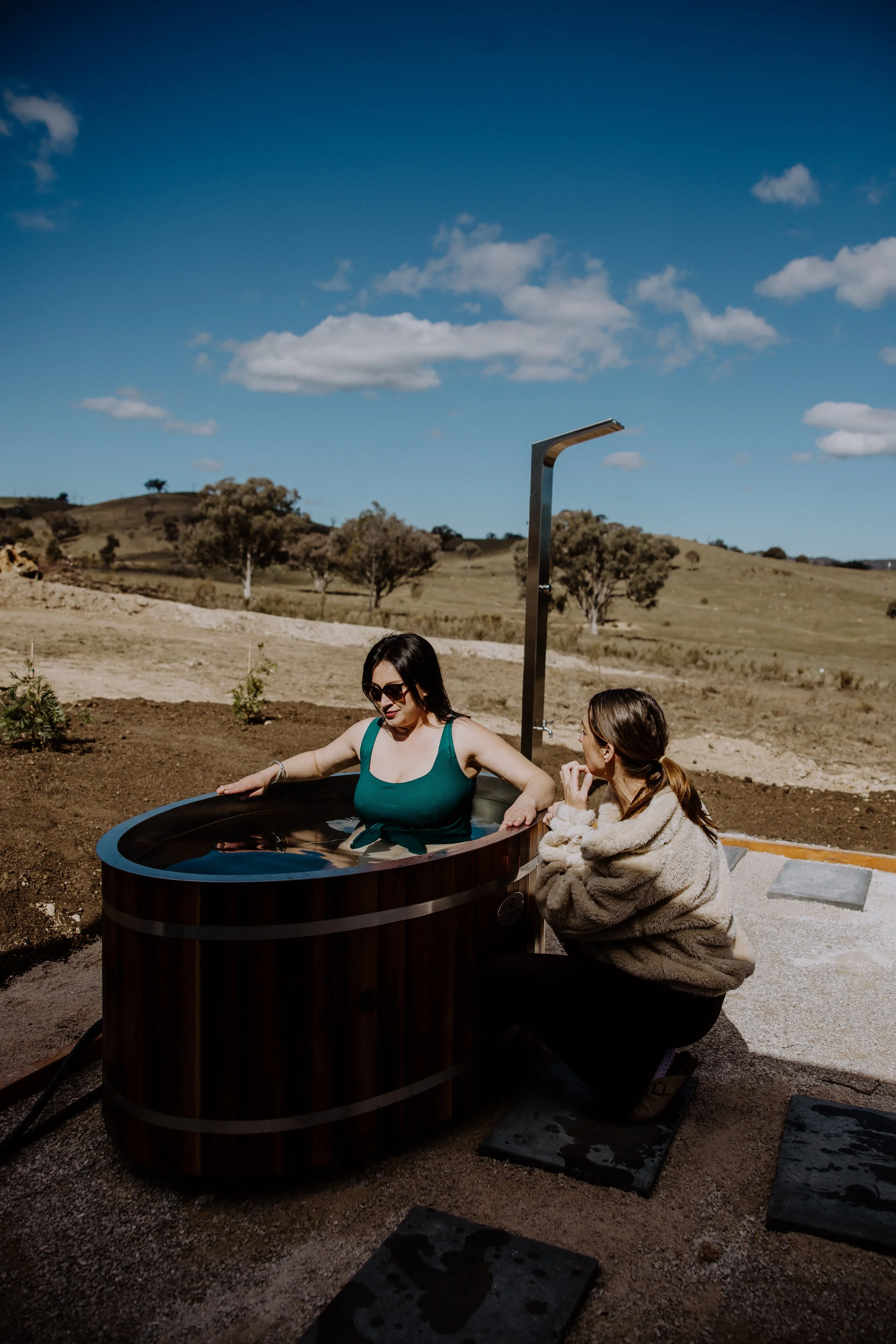 Two women, one sitting in a wooden hot tub outdoors on a sunny day, and another sitting next to it. The woman in the hot tub is wearing sunglasses and a teal swimsuit, while the other woman is wrapped in a beige fur coat. The background includes a rural landscape with trees, hills, and a partly cloudy blue sky.