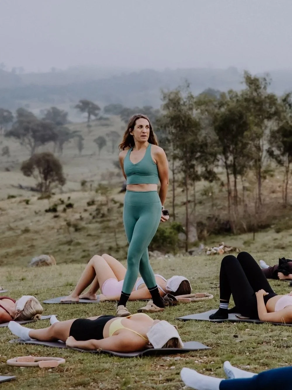 A woman in teal workout clothes leading an outdoor yoga class on a grassy hill, with several women lying on yoga mats practicing yoga, all wearing face masks.