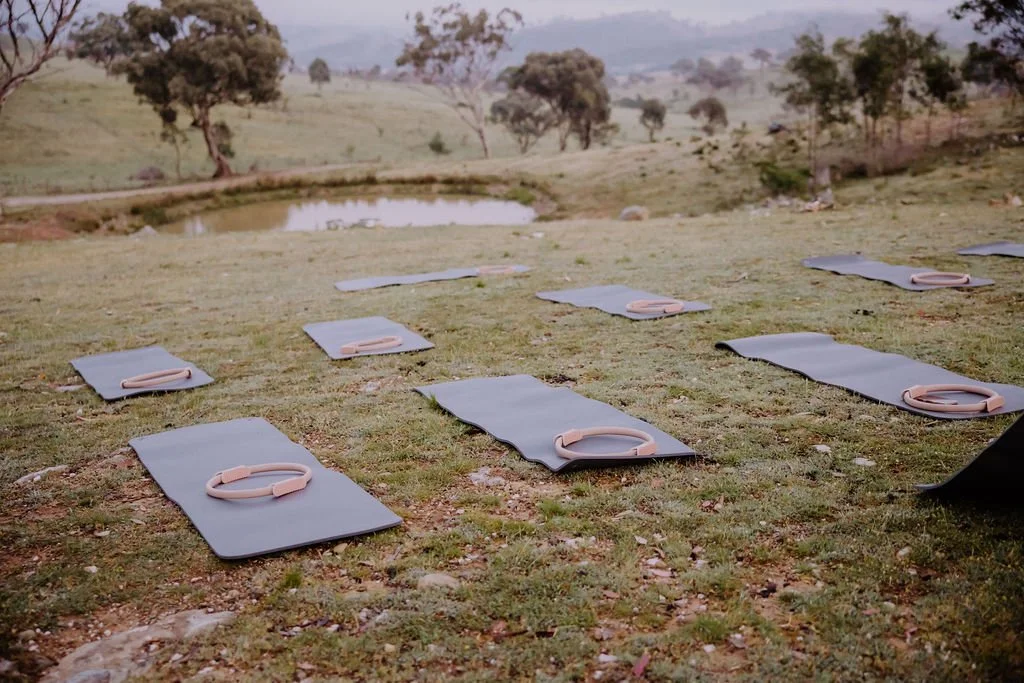 Yoga mats with straps arranged outdoors on grass near a small pond, with trees and rolling hills in the background.