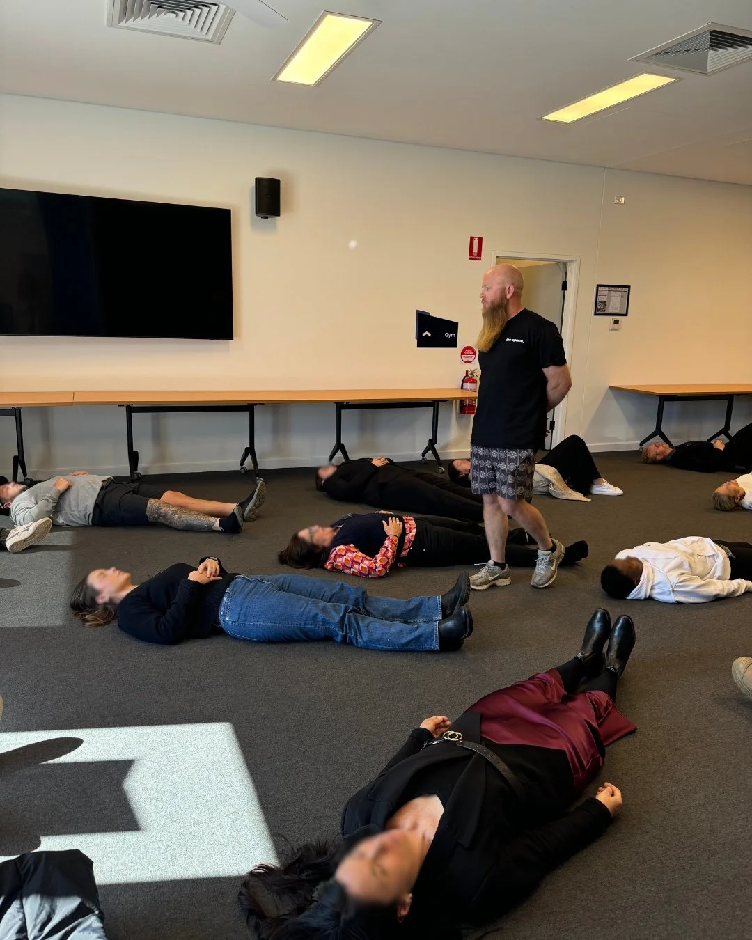 A group of people lying on the floor in a room, with their eyes closed or relaxed, while a man stands in the center leading a relaxation or meditation session. The room has a large television, tables, and wall-mounted speakers.