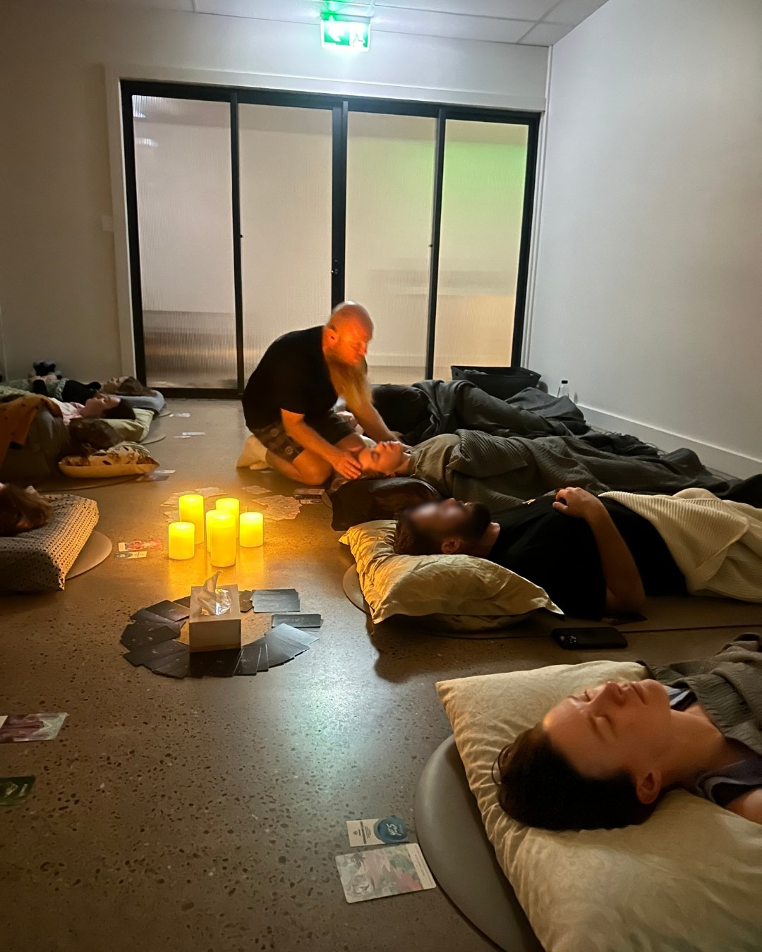 People lying on the floor, some resting with pillows, as a person appears to be performing a healing or spiritual practice on another person. There are candles and cards arranged on the floor, creating a calming atmosphere in a room with a sliding glass door and some scattered personal items.