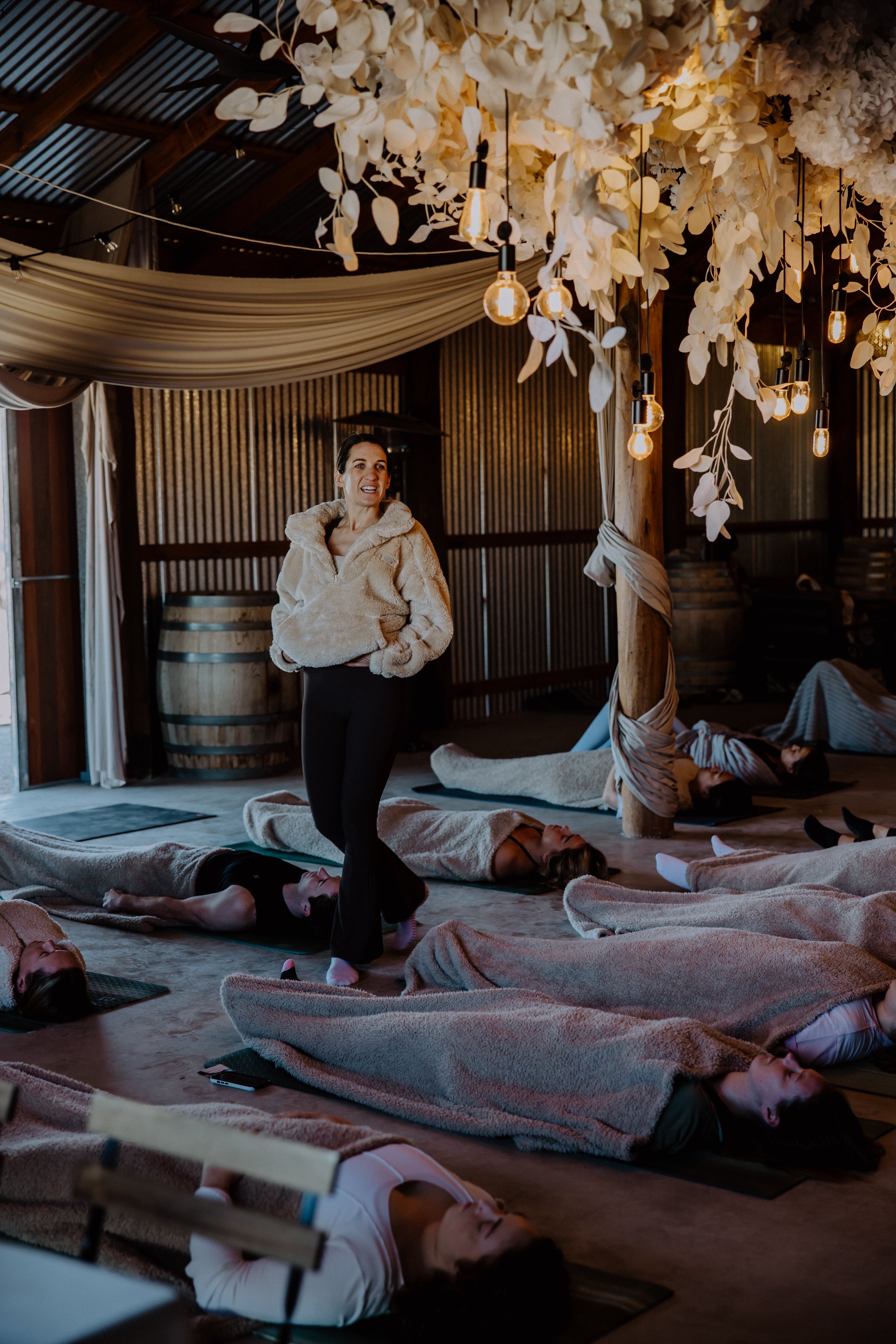 A woman leading a yoga or meditation class in a cozy, warmly lit studio with wooden walls and hanging lights, with several people lying on mats covered in blankets.