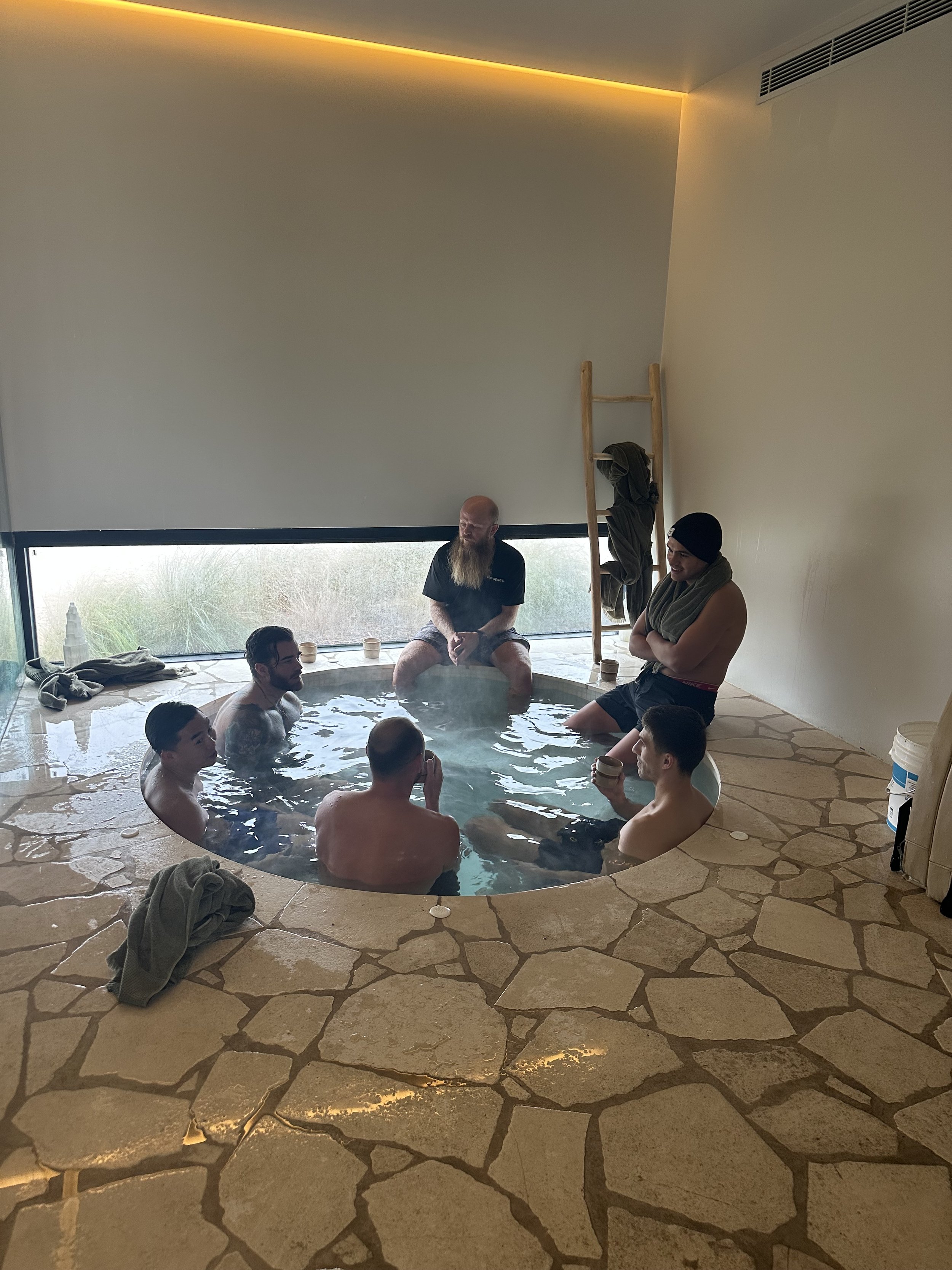 Group of people enjoying a hot tub inside a modern room with a stone floor and a large window.
