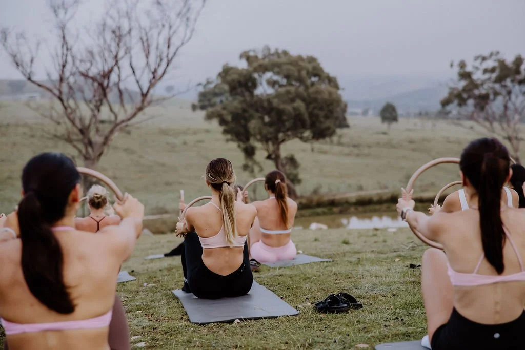 Group of women practicing outdoor yoga with hoops in a grassy field, trees, and a pond in the background.