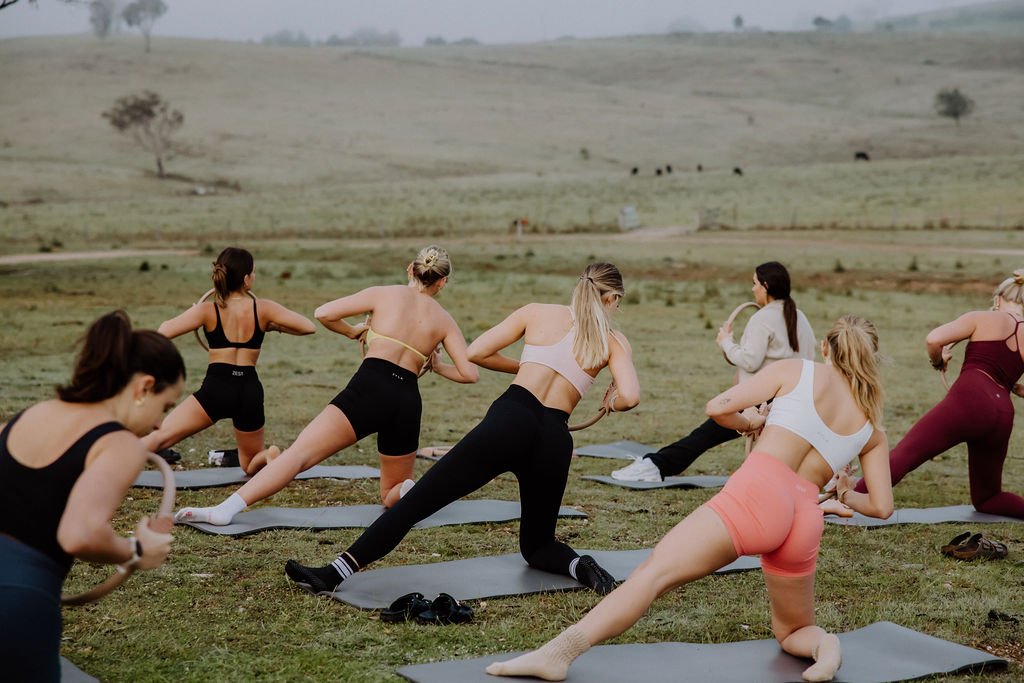 A group of women doing outdoor workout or yoga on mats in a grassy field.