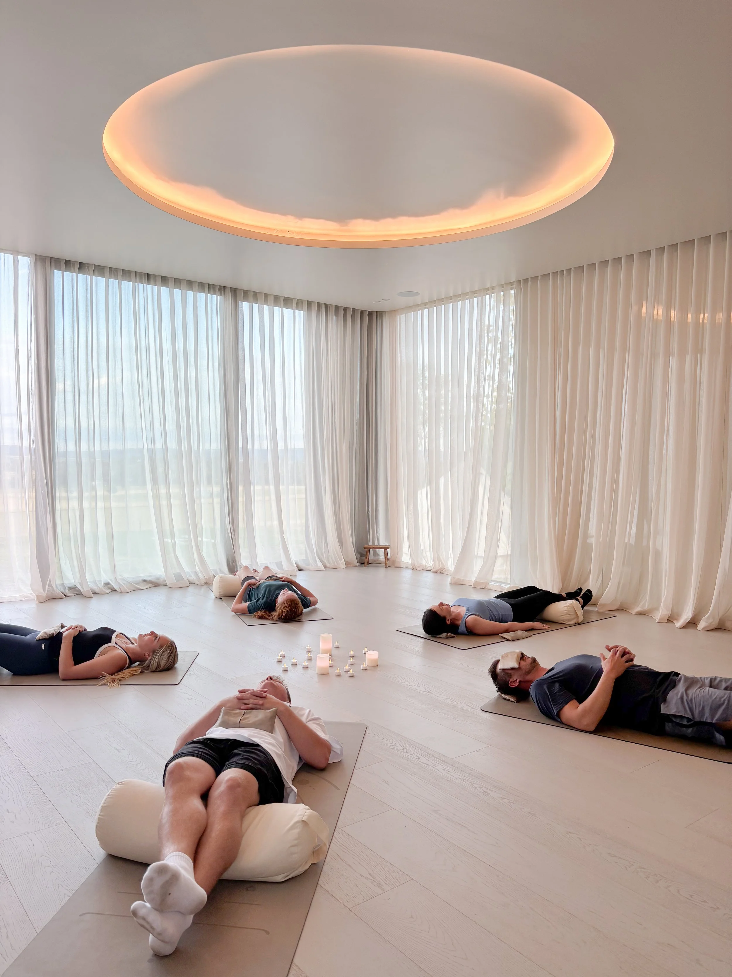 People participating in a relaxing yoga or meditation session in a bright, minimalistic room with large windows and sheer curtains, with candles on the floor.